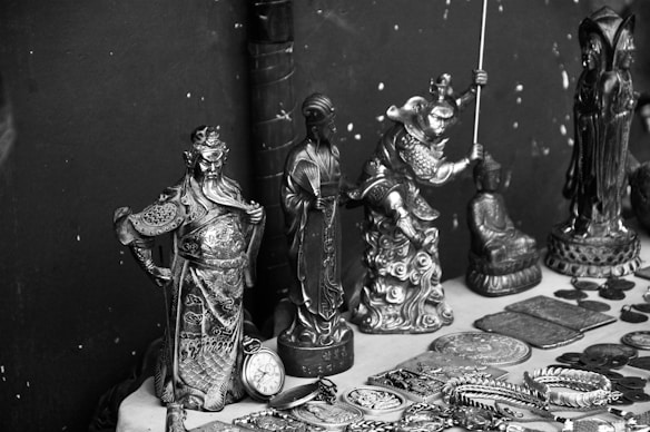 A collection of intricately designed metal figurines and objects displayed on a surface. The figurines depict various characters, some in dynamic poses, and are surrounded by several round coins and decorated plates. The items seem to be of cultural or historical significance and feature elaborate details in their craftsmanship.