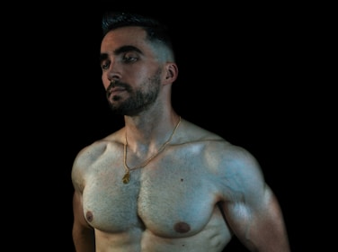 A shirtless man with well-defined muscles stands against a dark background. He has a beard, short hair, and wears a gold necklace. His expression is contemplative, and his gaze is directed to the side.
