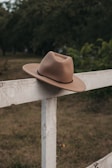 A brown cowboy hat rests atop a rustic white wooden fence in a serene outdoor setting. The background features lush greenery and grass, creating a peaceful countryside ambiance.