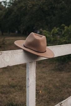 A brown cowboy hat rests atop a rustic white wooden fence in a serene outdoor setting. The background features lush greenery and grass, creating a peaceful countryside ambiance.