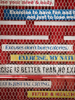 A metal fence with motivational fitness quotes written on colorful strips of tape in the background. The phrases include 'Exercise to have fun and be fit, not just to lose weight,' 'Excuses don't burn calories,' 'No exercise is better than no exercise,' 'Sweat is just fat crying,' and 'Fitter & healthier.' The strips are mostly white, blue, and yellow, placed on a red background.