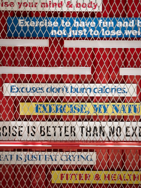 A metal fence with motivational fitness quotes written on colorful strips of tape in the background. The phrases include 'Exercise to have fun and be fit, not just to lose weight,' 'Excuses don't burn calories,' 'No exercise is better than no exercise,' 'Sweat is just fat crying,' and 'Fitter & healthier.' The strips are mostly white, blue, and yellow, placed on a red background.