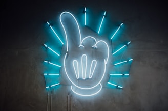 A neon sign featuring the shape of a hand with an extended index finger. The hand is white, while blue neon light rays radiate outward in a circular pattern, set against a dark gray wall.