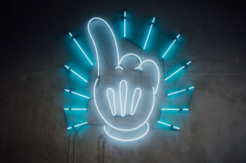 A neon sign featuring the shape of a hand with an extended index finger. The hand is white, while blue neon light rays radiate outward in a circular pattern, set against a dark gray wall.