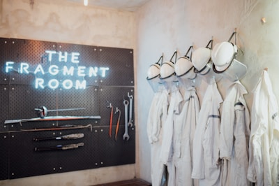 A wall display with the text 'THE FRAGMENT ROOM' illuminated in neon lights, accompanied by a pegboard featuring various tools such as hammers, wrenches, and sticks. On the right, a row of white protective garments and hard hats hang neatly, suggesting a workspace or activity area focused on safety and possibly destruction.