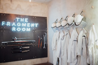 A wall display with the text 'THE FRAGMENT ROOM' illuminated in neon lights, accompanied by a pegboard featuring various tools such as hammers, wrenches, and sticks. On the right, a row of white protective garments and hard hats hang neatly, suggesting a workspace or activity area focused on safety and possibly destruction.