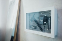 A framed display case mounted on a wall containing various black objects and tools. The items include a black strap with the label 'REMOVE BEFORE FLIGHT' and other items with 'ABOUT' branding. The case has a clean, modern design and the wall appears to be textured and painted in a light color. A mirror or reflective surface is leaning against the wall.