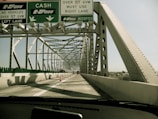 A vehicle is driving over a steel truss bridge with multiple lanes. Overhead signage indicates directions for cash and E-ZPass lanes, along with weight restrictions for vehicles. The bridge is constructed with large steel beams and has an open design that allows views of the sky and surrounding landscape.