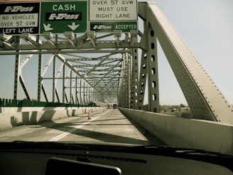 A vehicle is driving over a steel truss bridge with multiple lanes. Overhead signage indicates directions for cash and E-ZPass lanes, along with weight restrictions for vehicles. The bridge is constructed with large steel beams and has an open design that allows views of the sky and surrounding landscape.