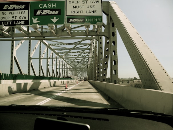 A vehicle is driving over a steel truss bridge with multiple lanes. Overhead signage indicates directions for cash and E-ZPass lanes, along with weight restrictions for vehicles. The bridge is constructed with large steel beams and has an open design that allows views of the sky and surrounding landscape.
