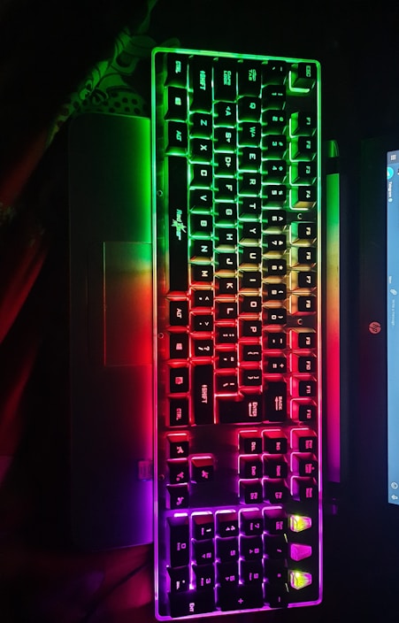 A mechanical keyboard with vibrant RGB backlighting is placed on a laptop. The keys are illuminated in a gradient of colors, ranging from green at the top to red, and then purple at the bottom, creating an eye-catching effect.