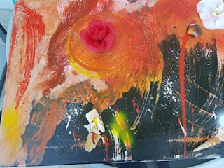 An abstract painting with a vibrant mix of colors, featuring a prominent red circular shape in the center. The paint is applied with bold strokes, combining red, orange, black, and yellow hues. A three-dimensional red flower is placed on the central circular shape. Scattered across the composition are white and tan seashells and textured pieces.