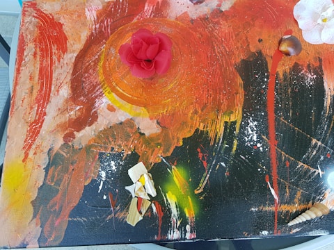 An abstract painting with a vibrant mix of colors, featuring a prominent red circular shape in the center. The paint is applied with bold strokes, combining red, orange, black, and yellow hues. A three-dimensional red flower is placed on the central circular shape. Scattered across the composition are white and tan seashells and textured pieces.
