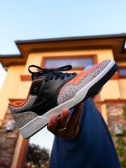 A hand is holding a stylish sneaker with a unique design featuring black leather, orange accents, and a patterned gray section. The background shows the exterior of a house with light-colored walls, stone detailing, and a clear sky.