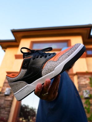 A hand is holding a stylish sneaker with a unique design featuring black leather, orange accents, and a patterned gray section. The background shows the exterior of a house with light-colored walls, stone detailing, and a clear sky.
