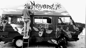 Two people are painting a van with elaborate and artistic designs. One person is on a stepladder painting the roof, while the other is on the ground working on the side panel. The van is covered in various artistic elements, including faces, animals, and abstract shapes.