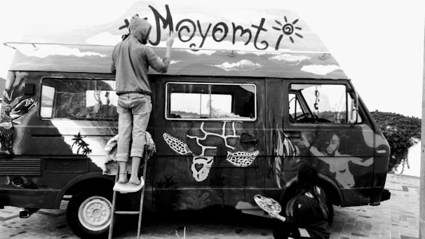 Two people are painting a van with elaborate and artistic designs. One person is on a stepladder painting the roof, while the other is on the ground working on the side panel. The van is covered in various artistic elements, including faces, animals, and abstract shapes.