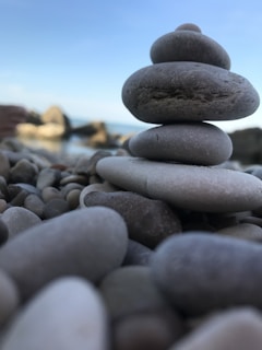 A stack of smooth, flat stones balanced on top of each other, set against a background of a rocky shoreline and a clear blue sky. The stones vary in size and create a sense of balance and tranquility.