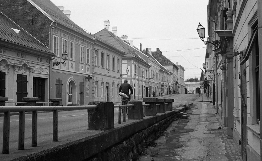 grayscale photo of man in black jacket standing on sidewalk near building