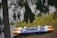 A blue and orange inflatable raft rests on the grassy edge of a body of water, partially obscured by the branches of a nearby tree.