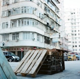 Wooden pallets are stacked in front of a multi-story residential building in an urban area. The building features numerous windows and air conditioning units. A black vehicle is parked nearby, with a yellow van and other traffic visible in the background. Signs with various text are attached to the building facade.
