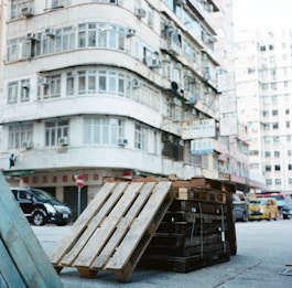 Wooden pallets are stacked in front of a multi-story residential building in an urban area. The building features numerous windows and air conditioning units. A black vehicle is parked nearby, with a yellow van and other traffic visible in the background. Signs with various text are attached to the building facade.