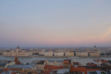 A panoramic view of a cityscape with a river running through it, flanked by historic buildings, including a prominent parliament building and a church with distinctive architectural features. The sky exhibits shades of pink and blue, suggesting a serene atmosphere during either sunrise or sunset.