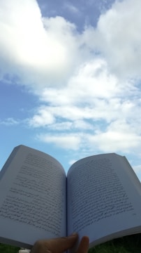A hand holds an open book with Arabic text against a backdrop of a clear blue sky with scattered white clouds. The book's pages are prominently displayed, capturing the texture and script.