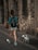 A person with a backpack is walking a small white dog on a leash along a stone-paved street. The setting includes an old stone wall, and the person is wearing a blue-patterned shirt, white shorts, and white sneakers.
