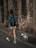 A person with a backpack is walking a small white dog on a leash along a stone-paved street. The setting includes an old stone wall, and the person is wearing a blue-patterned shirt, white shorts, and white sneakers.