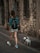 A person with a backpack is walking a small white dog on a leash along a stone-paved street. The setting includes an old stone wall, and the person is wearing a blue-patterned shirt, white shorts, and white sneakers.