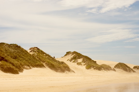 Sand dunes are covered with patches of green vegetation under a vast, partly cloudy sky. The sand is light beige, creating a warm and tranquil atmosphere. A person is visible in the distance on the dunes, adding a sense of scale to the landscape.