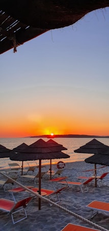 A serene beach scene at sunset with orange and red hues reflecting on the water. Several thatched umbrellas and orange lounge chairs are arranged neatly on the sandy shore, creating a calm and inviting atmosphere. The sky transitions from deep orange near the horizon to lighter shades of blue towards the top, with a sun slowly setting in the distance.