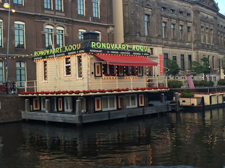 A picturesque canal scene featuring a houseboat adorned with string lights and vibrant red flowers. The boat's facade showcases large signage with green lettering. Adjacent to this is an ornate historical building with arched windows, providing an architectural contrast between the traditional and the modern atmosphere.