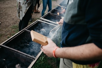 Several people stand around a row of large grills filled with charcoal, with smoke rising as flames flicker. One person holds a wooden item, possibly for handling food or charcoal, wearing casual clothing with a red wristband. The setting is outdoors on a grassy area.