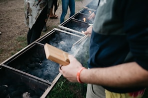 Several people stand around a row of large grills filled with charcoal, with smoke rising as flames flicker. One person holds a wooden item, possibly for handling food or charcoal, wearing casual clothing with a red wristband. The setting is outdoors on a grassy area.