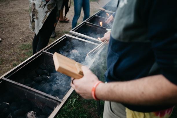 Several people stand around a row of large grills filled with charcoal, with smoke rising as flames flicker. One person holds a wooden item, possibly for handling food or charcoal, wearing casual clothing with a red wristband. The setting is outdoors on a grassy area.