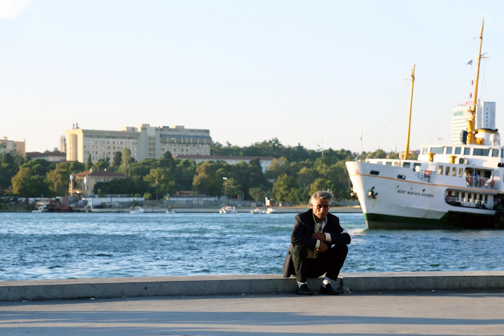 woman in black jacket and black pants sitting on gray concrete pavement near body of water