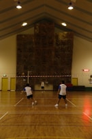 Three people are playing a game of badminton inside a sports hall with a high ceiling. The hall has a large climbing wall at the end and is lit by several overhead lights. The floor is wooden, and the players are wearing similar sporty attire.