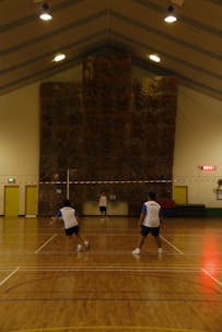 Three people are playing a game of badminton inside a sports hall with a high ceiling. The hall has a large climbing wall at the end and is lit by several overhead lights. The floor is wooden, and the players are wearing similar sporty attire.