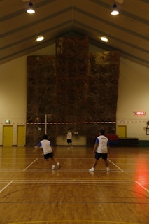 Three people are playing a game of badminton inside a sports hall with a high ceiling. The hall has a large climbing wall at the end and is lit by several overhead lights. The floor is wooden, and the players are wearing similar sporty attire.