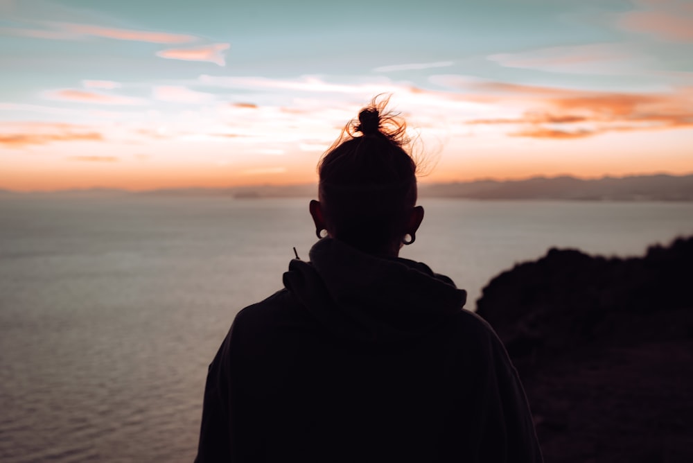 person in black hoodie standing near body of water during sunset