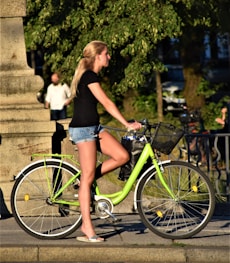 woman in black tank top and blue denim shorts standing beside green bicycle during daytime