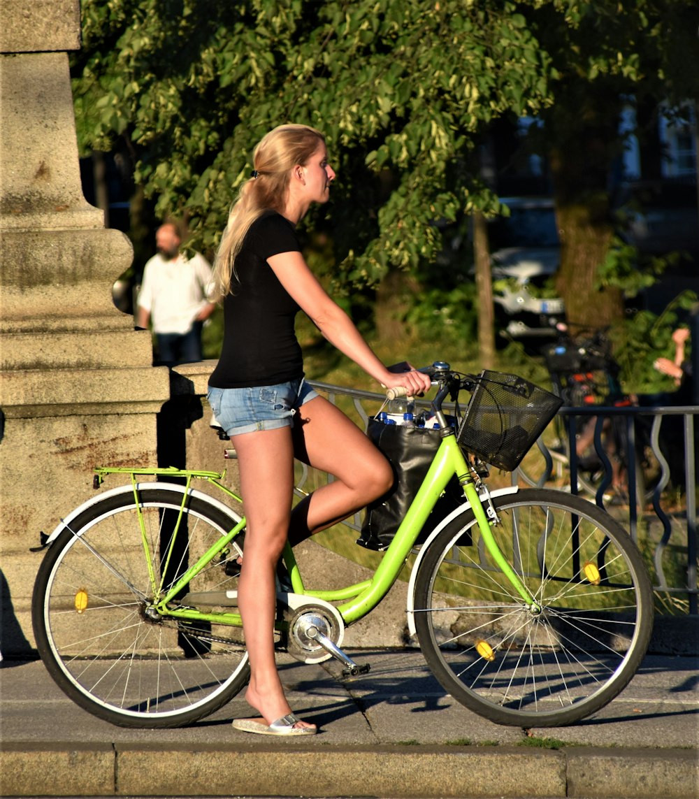 woman in black tank top and blue denim shorts standing beside green bicycle during daytime