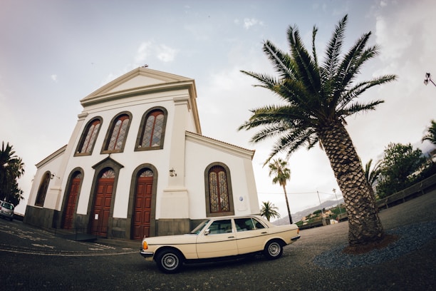 An old-fashioned car parked outside a small church with peeling white paint under a cloudy sky.