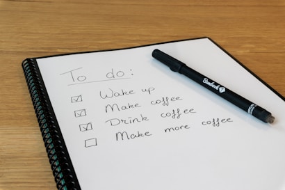 A spiral-bound notebook with a to-do list written in neat handwriting. The tasks include waking up, making coffee, drinking coffee, and making more coffee, with checkboxes beside them. Three tasks are checked off. A black pen is resting on the notebook, and the background is a wooden surface.