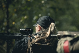 A person with long blonde hair wearing a black cap and backpack is holding a rifle with a scope. The setting appears to be outdoors in a forested area, with blurred greenery in the background.