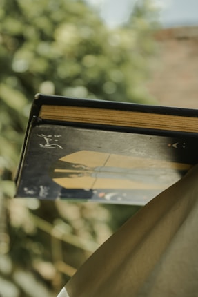 A book with a black cover and gold pages is partially wrapped in fabric, resting against a blurred natural background. The cover features a graphic design with subtle, artistic elements.