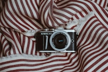 A vintage-style camera placed on a piece of fabric with red and white stripes. The lens is prominently visible, and the camera's brand name is displayed above the lens. The fabric appears soft with fringed edges, creating a cozy and textured background.