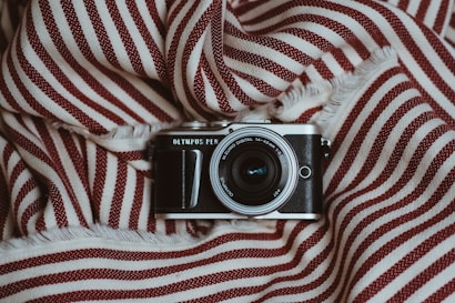 A vintage-style camera placed on a piece of fabric with red and white stripes. The lens is prominently visible, and the camera's brand name is displayed above the lens. The fabric appears soft with fringed edges, creating a cozy and textured background.
